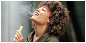 Happy woman smiling while smoking a vape outdoors. She has curly hair and she is mixed race. She is holding the disposable vape and exhaling smoke with her head tilted.