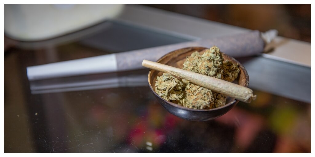 Close up of a joint placed on top of raw marijuana buds in a small bowl and another joint in the background