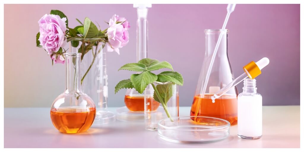 a setup of bottles with orange liquid, laboratory utensils, a peony flower and a sprig of mint